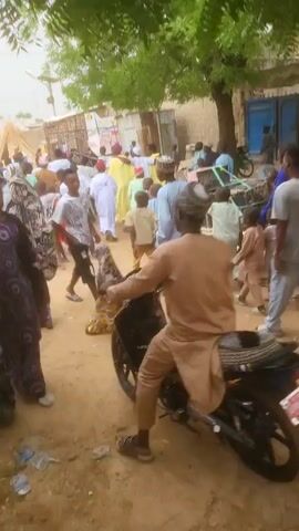 The video shows a busy street scene with many people, including adults and children. Most are walking, but one man is riding a motorcycle, and a boy is on a bicycle. Several individuals wear traditional clothing, and some women have headscarves. The ground appears to be dirt, littered with plastic bottles and other debris. There are buildings in the background, including one with a blue door, and trees providing shade. The camera moves slightly, panning across the scene, and there's music playing in the background.