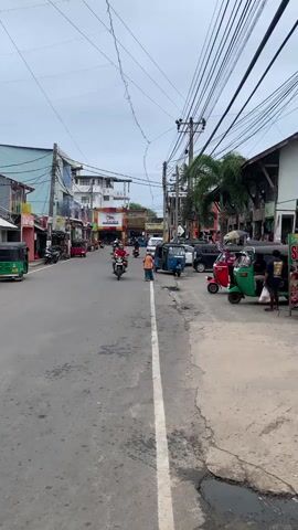 The video shows a street scene with several buildings lining both sides of the road. The road is paved and has a white line dividing the lanes. There are several vehicles on the road including motorcycles, tuk-tuks, and a van. People are seen riding the motorcycles and walking on the sidewalk. The buildings appear to be shops and businesses. There are overhead power lines with wires crisscrossing the street. The sky is cloudy. The video appears to be filmed from a moving vehicle.