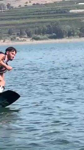 Here is a description of the video:

The video features a man wakeboarding on a body of water, possibly a lake or a wide river. He is wearing a dark-colored life vest and dark shorts. His posture is bent at the waist, with his arms holding onto a rope attached to a boat that is pulling him. He is positioned sideways on a dark wakeboard, creating a spray of water as he moves.

The backdrop shows a hilly landscape with vegetation and some buildings, indicating a populated area along the shore. The sky is bright, and the water reflects a dark color, contrasting with the white foam created by the wakeboard. The camera follows the man as he moves, keeping him centered in the frame and showing his movements and the water rippling behind him.