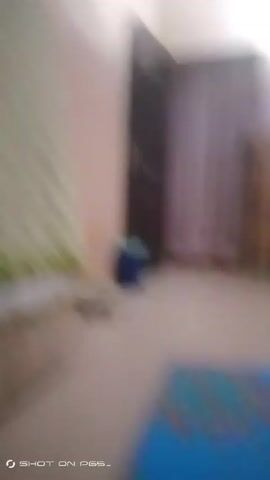 The video shows a blurry, shaky pan of a room. There is a bed with a green blanket to the left, a door and a curtain on the right, and a refrigerator further in the room. The walls are painted in two tones, and there is a window at the back.  The overall image quality is poor due to blur and camera shake.