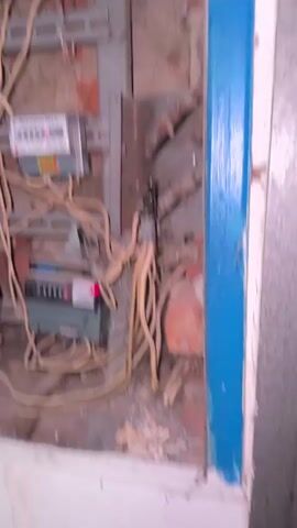 The video shows an electrical panel with multiple electricity meters and circuit breakers. The panel is located in a brick wall. The wiring appears to be old and disorganized. The video starts with a wide shot of the panel, then zooms in on the meters and breakers. A hand is seen opening a cover to reveal more circuit breakers. The scene appears to be in a storage room or utility closet.