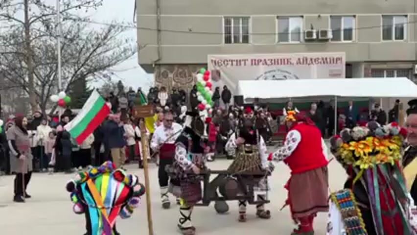 Here’s a description of the video:

The video shows a festive gathering in what appears to be a town square in Bulgaria. The atmosphere seems lively and celebratory, with many people dressed in traditional Bulgarian clothing and costumes. There are dancers performing, some carrying or manipulating a wooden farming tool that resembles a plow. 

A man in religious attire, a priest perhaps, is also part of the group, blessing the event with green branches. A crowd of onlookers, including children and adults, is gathered to watch the performance, with some holding Bulgarian flags and colorful balloons. 

In the background, there's a building with a banner that reads "ЧЕСТИТ ПРАЗНИК АЙДЕМИР!" (Happy Holiday Aidemir!). There's also a tent set up, and the overall setting suggests a public celebration or festival.