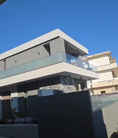 The video shows a contemporary, two-story house with a gray facade and white accents. The house features a balcony with glass railings on the upper level and a stone-clad base on the lower level. A dark gray wall encloses the property, and landscaping includes small palm trees and planters. The sky is clear and blue.