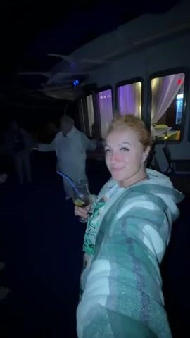 A woman with reddish-blonde hair, wearing a green and white plaid jacket over a white shirt, holds a drink with a blue straw and smiles at the camera. She is on a dark-colored deck, possibly on a boat, with large windows in the background that are illuminated with purple light. Several other people are visible in the background, some dancing. One person in a white outfit appears to be dancing in the middle ground. The lighting is dim, with some colored lights adding to the atmosphere.