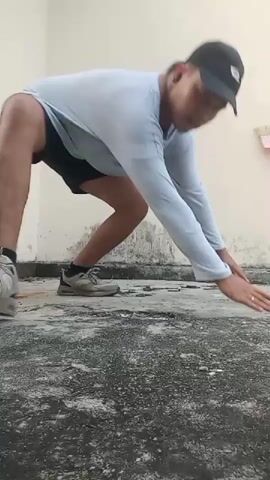 The video shows a person performing push-ups on a textured concrete floor. The person is wearing a light blue long-sleeved shirt and black shorts. They are positioned in a plank pose with their hands on the ground, shoulder-width apart. The person then lowers their body towards the ground, bending their elbows, and then pushes back up to the starting position. This motion is repeated several times. The background consists of a plain wall and the floor has some dark stains and debris. The lighting appears to be natural daylight.
