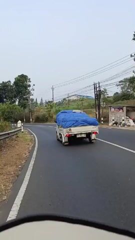 The video is recorded from the perspective of someone on a motorcycle, following a small white truck with a blue tarp covering its cargo. The truck is traveling on a two-lane asphalt road that curves to the left. On the left side of the road, there is a grassy shoulder with a guardrail and a line of white posts with black tops. Trees and vegetation are visible on both sides of the road. In the distance, there are power lines and poles. A motorcycle with a rider is also seen in the distance on the right side of the road. Further down the road, another truck, which appears to be a larger transport truck, is parked on the right shoulder. The sky is overcast, suggesting it might be early morning or late afternoon.