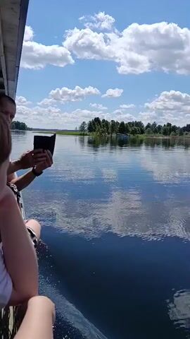 A person on a boat holds a dark object, possibly a tablet or book, and waves. They are standing next to a railing on the side of the boat, facing the camera and a wide body of water. The water is calm and reflects the bright blue sky with scattered white clouds. Trees and a distant shoreline are visible on the other side of the water. Another person's arm rests on the railing in the foreground. The sun appears to be shining brightly.