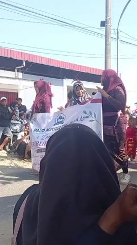 A group of people, mostly women wearing hijabs and traditional clothing, are participating in a parade. They are carrying a banner that reads "REPUBLIK INDONESIA 78 AGUSTUS 1945". Some people are walking, while others are riding on a decorated vehicle that appears to be a cart or float. There are spectators lining the street, and some of them are holding up phones to record the event. The environment appears to be a street with buildings on one side. The overall atmosphere seems festive.