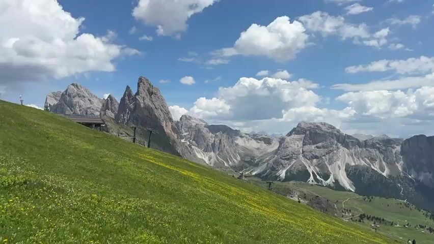 This media file captures a breathtaking mountain landscape under a bright, cloudy sky. The foreground is dominated by a lush, green slope dotted with small yellow flowers, creating a vibrant and natural texture. A well-maintained path, likely for hiking or skiing, is visible winding its way down the slope and into the valley below.

In the mid-ground, the dramatic peaks of the Dolomites rise majestically. These are rocky, jagged mountains with steep cliffs and visible patches of snow, suggesting a high altitude. A ski lift system, with its characteristic towers and cables, is subtly integrated into the scene, hinting at recreational activities. A small building, possibly a mountain restaurant or a station for the ski lift, is perched on the hillside.

The background reveals a vast mountain range stretching into the distance, with layers of peaks fading into a hazy blue. The sky is a brilliant blue, punctuated by fluffy white cumulus clouds that cast shadows on the mountains, adding depth and drama to the scenery.

The time of day appears to be daytime, likely late morning or early afternoon, given the bright sunlight and the clarity of the scene. The weather is pleasant, with a mix of sun and clouds, typical for a summer day in the Alps. The overall impression is one of serene natural beauty and a popular destination for outdoor enthusiasts. The location cues, such as the distinctive rock formations and the presence of ski infrastructure, strongly suggest the Dolomites in Italy, specifically the region around Santa Cristina Gherdëina. The scene evokes feelings of awe, tranquility, and the exhilaration of being in a grand natural environment. There is no visible text in the media file.