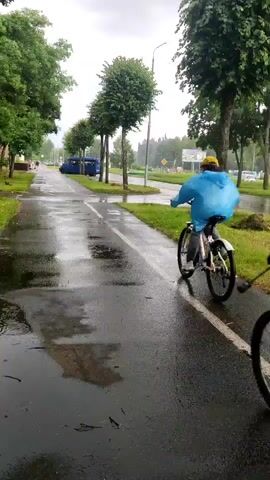 Two individuals are riding bicycles on a wet paved path alongside a grassy area and trees. The path is wet from rain, with puddles and reflections visible. Both cyclists are moving away from the camera. The person in the foreground is wearing a pink hooded jacket and a backpack. The person in the background is wearing a blue hooded rain poncho and a yellow helmet. The environment appears to be an urban or suburban setting with buildings and streetlights visible in the distance. The sky is overcast, indicating continued rain or recent rainfall.