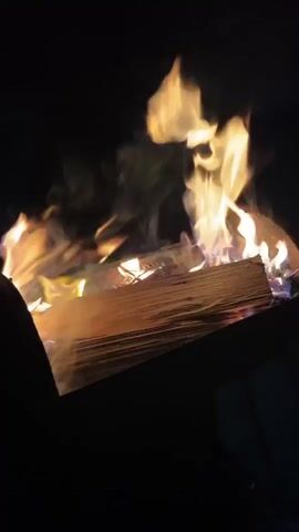 The video displays a close-up shot of logs burning in a fire pit at night. The flames are orange, yellow, and white, with wisps of smoke rising into the darkness. The logs appear to be split firewood, stacked horizontally. The fire is the primary light source, illuminating the wood and casting a warm glow. The background is entirely black, indicating a dark environment. No people are visible in the frame.