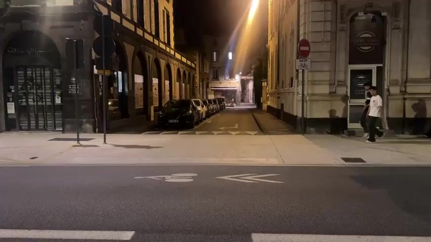 The video shows a street scene at night. The street is lined with buildings on both sides. Cars are parked along the street on the left side of the frame. There is a bicycle lane marked on the road in the foreground. A white van is driving past on the right side of the frame, its headlights and taillights are visible. A person is walking on the sidewalk on the right side of the frame. Another person is visible further down the sidewalk. The street is illuminated by streetlights. There are traffic signs and street furniture visible.