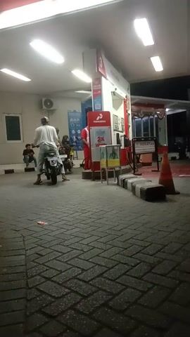 A gas station at night with several people and motorcycles. A man in a white shirt and helmet is sitting on a motorcycle. Another person is standing behind him. The gas pumps are red and white. The ground is paved with bricks. There are fluorescent lights overhead.