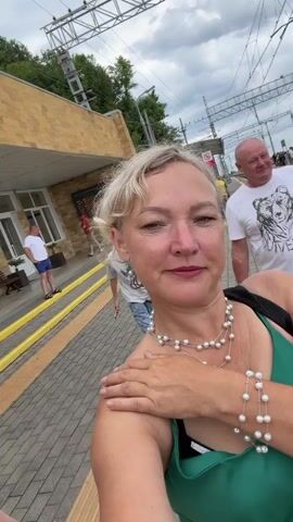 The video shows a woman in a green top and pearl necklace taking a selfie at a train station. In the background, other people are visible, including a woman in a white shirt and a boy with red hair. The station building is made of yellow brick, and there are train tracks and overhead wires in the distance. The sky is cloudy.