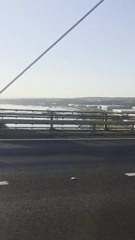 The video shows a view from a vehicle driving on a bridge. The bridge has multiple lanes and is suspended by cables. The sky is clear and blue. On the bridge, there are several other vehicles, including a white van and trucks. The scenery beyond the bridge appears to be a city or town with buildings and some greenery. The video is shot from a slightly elevated perspective, giving a sense of the height of the bridge.