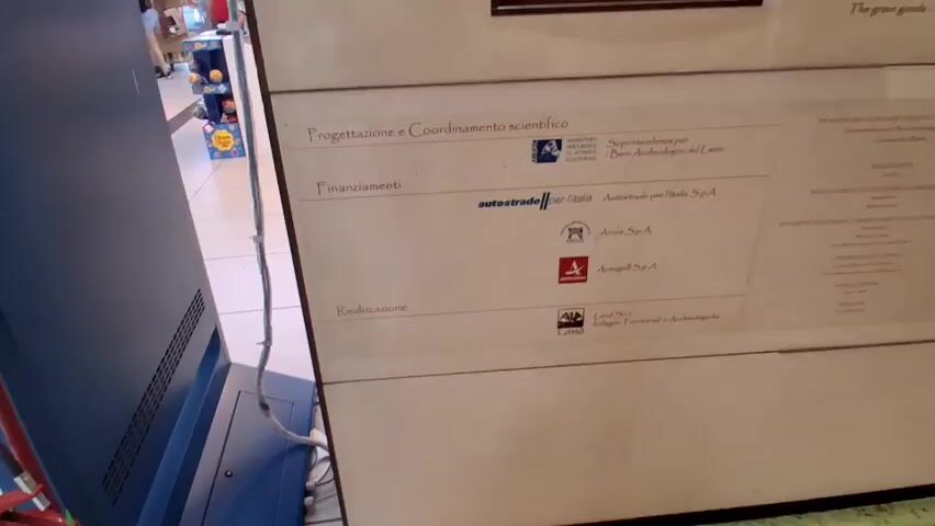 A wide-angle shot focuses on a white informational panel. The panel is divided into sections with text and logos. The top section, titled "Progettazione e Coordinamento scientifico," lists "Ministero per i Beni Culturali e Ambientali" and "Soprintendenza per i Beni Archeologici del Lazio" with their respective logos. The next section, "Finanziamenti," features the "Autostrade per l'Italia" logo and "Autostrade per l'Italia SpA," followed by the logos and names of "Arca SpA" and "Anas SpA." The third section, "Realizzazione," shows the "Land Sci" logo and "Land Sci - Servizi Territoriali e Archeologici." To the right, another column of text lists similar information, including "Direzione Progettazione Scientifica e Coordinamento," "Realizzazione," "Progettazione Architettonica e Direzione Lavori," "Redazione Testi e Progettazione Allestimenti e Trine," "Comunicazione Grafica," and "Modellistica." The panel is mounted on a darker background, possibly a wall, and a portion of a blue electronic device with cables is visible on the left side of the frame. The environment appears to be indoors, possibly a museum or public exhibition space, with hints of displays or merchandise in the blurred background.