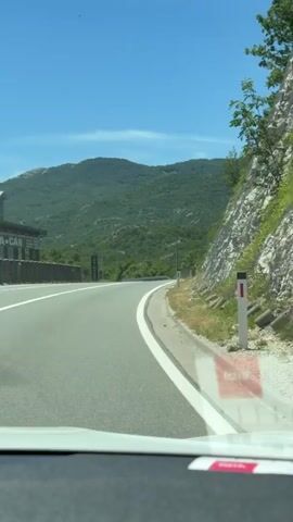 The video shows a first-person perspective from inside a vehicle driving on a paved road. The road curves to the right. On the left side of the road, there are some signs and a fence. On the right side of the road, there is a steep, rocky embankment with some vegetation. In the background, there are green, tree-covered mountains under a clear blue sky. A billboard advertising a car is visible on the right side of the road. The dash of the car is visible in the foreground.