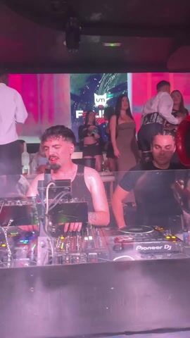 The video shows two DJs mixing music in a nightclub. The DJs stand behind a Pioneer DJ mixer and turntables, adjusting knobs and faders, and manipulating records. The club is dimly lit, with red and pink lights flashing. Behind the DJs, a video screen displays the club’s logo. People are dancing, drinking, and socializing on the dance floor. The camera moves slightly, focusing on the DJs as they work.