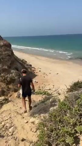 The video shows a person walking down a sandy path towards a beach. The person is male, wearing a black t-shirt and shorts, and a baseball cap. He is carrying a white object that appears to be a surfboard. The path leads from a cliff down to a sandy beach with turquoise water. The sky is clear and blue. Some vegetation is visible along the path. The person appears to be walking alone.