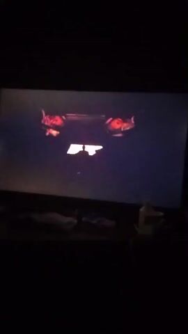 A dimly lit room shows a television screen displaying a scene from a video game or movie. The screen is dominated by a dark purple and black background, with a bright orange and red opening in the center, resembling a doorway or portal. A silhouetted figure stands in the center of the opening, facing away from the viewer. Two large, claw-like or horn-like shapes, glowing with orange light, extend from the top edges of the screen towards the center. The bottom edge of the television is visible, with a dark surface below it. A few indistinct objects are on the surface, including what appears to be a white bottle on the right. The lighting is dark, with the television screen being the primary light source in the room.