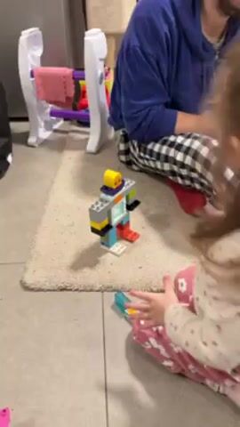 Two children are sitting on a rug, facing a robot made of Duplo blocks. The robot has a yellow head with a smiley face, a gray body with a white door, and blue and red legs with white feet. A little girl wearing pink pajamas points at the robot. Another child in a blue sweatshirt and checkered pajamas is sitting near the rug. Behind them is a white toy rack. The rug is tan and the floor is light gray tile. A gurgling or crying sound can be heard.