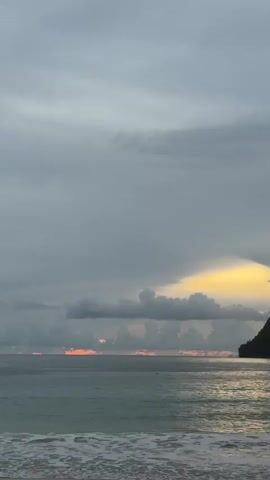 A vertical video shows a body of water with waves lapping at the shore, under a cloudy sky. A dark silhouette of a landmass is visible on the right side of the frame, just above the water. The sun appears to be setting or has recently set, as there are streaks of orange and yellow light visible on the horizon between the clouds. The water is a muted blue-green, and the waves are white. The sky is filled with various shades of gray clouds, with a bright patch of yellow light peeking through on the right. No people or specific objects are clearly discernible in the water or on the landmass. The overall mood is serene and perhaps a bit somber due to the overcast sky.