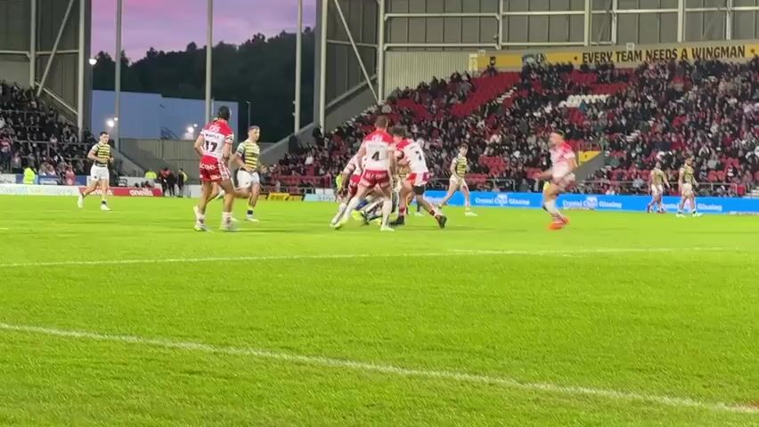 The video shows a rugby game being played on a green grass field in a stadium. Spectators are seated in the stands, which are mostly red. The players are wearing red and white or yellow and black striped jerseys. The players in red and white are clustered together, some on the ground, suggesting a tackle or scuffle. A player in a red and white jersey with the number 5 on his back is visible in the foreground. The sky is visible in the background with sunset colors. The overall scene is a live sports event.