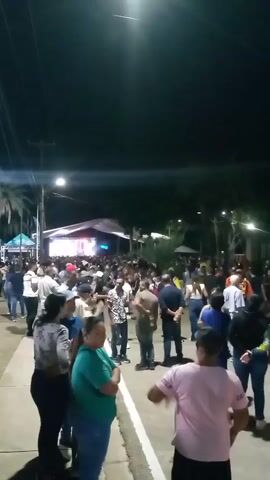 A nighttime outdoor event is depicted with a crowd gathered around a stage. The stage is illuminated with bright lights and a large screen displaying visuals. People are standing and milling about in the foreground and middle ground, facing the stage. Some are dressed casually, while others appear to be dressed for a festive occasion. The environment suggests a town square or public gathering space, with some palm trees visible in the background and streetlights illuminating the area. The overall atmosphere appears to be lively and engaged.