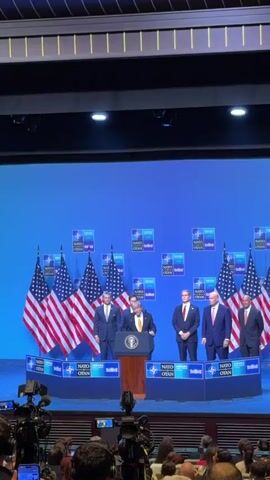 The video shows a group of men on a stage in front of a blue backdrop with numerous NATO logos. In the center, a man wearing a dark suit and a yellow tie stands behind a podium with the Presidential Seal of the United States. He is speaking. To his left, there are several American flags. To his right, there are other men in suits standing. The stage is elevated, and the audience is visible in the foreground, with cameras and other recording equipment present. The overall setting appears to be a press conference or formal event associated with NATO.
