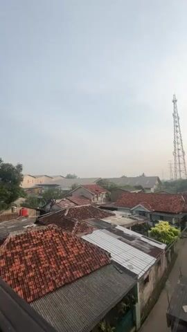 A video pans over a hazy residential area with a tall cell tower visible in the distance. Rooftops of houses, some with orange tile and others with corrugated metal, are prominent. A narrow road winds through the neighborhood. Trees and power lines are scattered throughout the scene. The sun is visible in the sky, creating a soft glow. The overall atmosphere suggests it is either early morning or late afternoon due to the haze and the position of the sun.