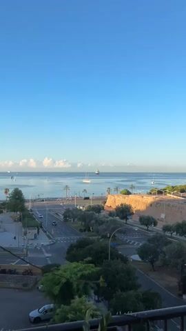 The video shows a panoramic view of a coastal city on a clear, sunny day. In the foreground, a balcony railing is visible with two green chairs. Beyond the balcony, a street runs parallel to the coastline, with cars driving on it. Trees line the street and a fortified wall. In the distance, the sea is calm with several sailboats and larger ships. A few fluffy white clouds are scattered across the bright blue sky. The scene appears to be from an elevated vantage point, likely a hotel room or apartment overlooking the city and the sea.