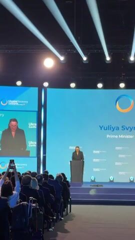 The video shows a woman speaking at a podium on a stage. She is wearing a black blazer and glasses. Behind her, a large screen displays the text "Ukraine Recovery Conference" and her name, "Yuliya Svyrydenko Prime Minister". The stage is lit with spotlights. In the foreground, the backs of people's heads are visible as they are seated in an audience, some are holding up phones to record the event. The overall atmosphere is that of a formal conference or presentation.