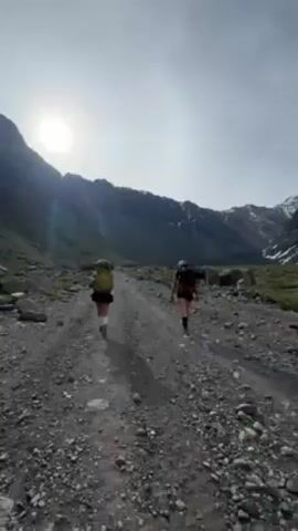 The video shows two people hiking on a rocky trail towards snow-capped mountains. The hikers are wearing backpacks and shorts. The trail is surrounded by rocks and sparse vegetation. The sky is bright and there are patches of snow on the mountain peaks. The camera follows the hikers from behind, and then pans to show the mountain scenery.