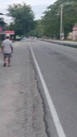 The video shows a slow-moving perspective, likely from a vehicle or camera operator walking alongside a road. The road is paved with a white line marking the edge on the left. There are trees and some buildings visible in the background. At one point, an elderly man wearing a light-colored shirt and shorts walks away from the camera on the dirt shoulder of the road. The sky appears overcast, and the overall lighting suggests it may be early morning or late afternoon. The video is shaky, indicating movement.