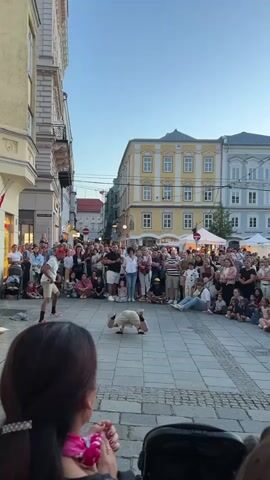 A street performance is taking place on a cobblestone street in front of historical buildings. A large crowd of spectators is gathered, some seated and others standing, observing the performance. In the center of the street, a performer is doing a breakdancing move, with one leg extended upwards and the other bent, supporting their body. Another performer, wearing a red cap and white shirt, stands to the left of the breakdancer, seemingly interacting with the crowd. The buildings are multi-story structures with architectural details typical of European cities. The sky is clear and blue, indicating it is daytime. The overall scene depicts a lively public gathering with an emphasis on street entertainment.