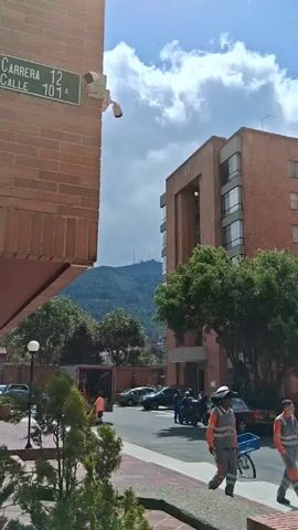 The video shows an urban scene with a mix of residential buildings, trees, and street activity. The buildings are primarily made of red brick. A street sign indicates "Carrera 12, Calle 107A." There's a security camera mounted on the corner of one building. In the distance, a mountain can be seen under a sky with scattered clouds. Several people are working on or around a truck, wearing orange safety vests. A yellow taxi, with license plate "WMM 459", drives past. The taxi has "SERVICIO PUBLICO" written on its side. A person in a similar orange safety vest walks along the sidewalk. There are other parked cars and motorcycles along the street. The overall environment is a mix of urban and natural elements.