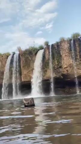 The video displays a waterfall with multiple streams cascading over a rocky ledge into a pool of water. The sky is partly cloudy. A medium-sized rock is visible in the water in the foreground, and some vegetation lines the top of the ledge. The camera appears to be slowly panning or zooming in closer to the waterfall.
