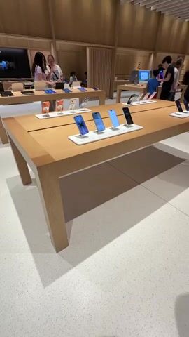 The video shows the interior of what appears to be an Apple Store. In the foreground, a long, light-colored wooden table displays several iPhones on stands. The phones are angled to show their screens. The phones have screens showing different images and apps. Behind this table, another similar wooden table is visible, also displaying iPhones. Further back, other tables and displays are present, along with a few people browsing or interacting with the products. The environment is clean and well-lit, with wooden accents and a speckled white floor. The overall atmosphere suggests a retail environment focused on technology products.
