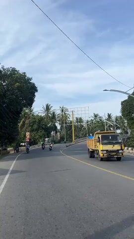 A person is filming from the perspective of a moving vehicle on a paved road with yellow lane markings. On the left side of the road, there are trees, a billboard with a yellow pole, and several people riding motorcycles. Ahead, the road curves to the right. On the right side of the road, there is a yellow truck, a person riding a motorcycle, and palm trees. The sky is blue with white clouds.