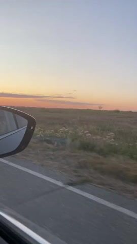 The video shows a view from inside a car. The camera is positioned to capture the side mirror on the left and the landscape outside on the right. The side mirror reflects the interior of the car, including a person who appears to be the driver or a passenger. Outside the car, a road with a white line marking runs horizontally across the frame. Beyond the road, there is a grassy field under a sky with a gradient of colors, transitioning from a light blue at the top to warm orange and pink hues near the horizon, indicating a sunset. The sun itself is visible low on the horizon, partially obscured by atmospheric haze. The field appears dry and stretches towards distant, low hills. The video is brief and shows the car moving forward, indicated by the blurred motion of the road and the field.