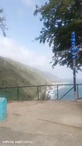 A video shows a scenic view of a coastline with a prominent cross in the foreground. The cross, which is decorated with blue and silver accents, stands to the right of the frame. Behind it, a lush green hillside slopes down towards the turquoise ocean. The sea is calm with small waves visible near the shore. A road can be seen winding along the coast. The sky is bright and clear with a few scattered clouds. A large tree with dark green leaves partially obscures the upper right portion of the frame. The video is shot from an elevated vantage point, suggesting it was taken from a cliff or overlook. A fence runs horizontally across the middle of the frame, separating the foreground from the view of the coastline. The overall atmosphere is serene and picturesque.