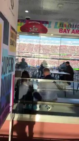 The video shows the interior of a VIP lounge overlooking a crowded stadium. People are seated at tables or standing, some looking out at the stadium. There are televisions on the walls, and a set of stairs leading to another level. The lounge has decorated pillars with colorful designs. The video shows a view of the stadium through the glass windows, as well as parts of the interior of the lounge.