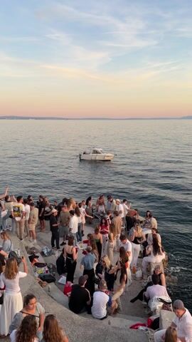 A high-angle wide shot depicts a gathering of approximately 60 people on a concrete pier or quay, adjacent to a body of water. The majority of individuals are standing or sitting in small clusters, while a few are taking photographs with their phones. The general attire suggests a casual, possibly summer, social event. In the background, a small white motorboat is anchored in the water, and the horizon line reveals distant land masses under a sky exhibiting hues of blue and pink from what appears to be a setting sun. The overall atmosphere seems relaxed and social.