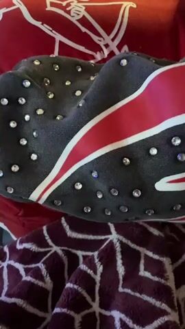 The video shows a close-up of several items of clothing. A gray sweatshirt with a red and white stripe and small, round, reflective embellishments is in the center. Below it is a maroon blanket with a geometric pattern. Above the sweatshirt is a red shirt with a white logo. The items appear to be on a bed or similar surface. The camera remains stationary throughout the video.