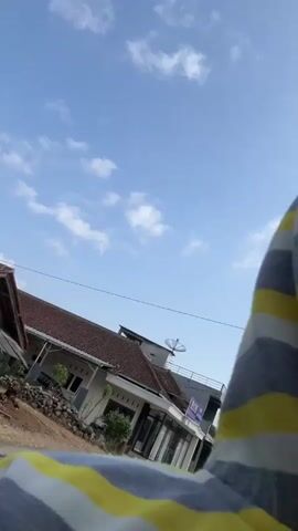 The video shows a low-angle view of a bright blue sky with scattered white clouds. In the foreground, a person's torso and shoulder are visible, wearing a striped shirt with alternating colors of white, gray, and yellow. The background features lush green trees with some houses and buildings interspersed. A street sign is visible on the left side of the frame. The camera appears to be moving, creating a slight blur and a sense of motion. The overall impression is that of an outdoor scene, possibly a leisurely drive or walk on a sunny day.