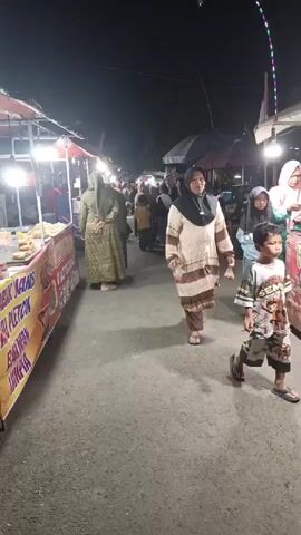 The video captures a nighttime scene at an outdoor market or fair. A young boy, wearing a dark t-shirt and a sarong, walks away from the camera down a paved walkway. To his left, a food stall is visible with bright lights and banners displaying text. Several people are browsing the stalls and walking along the path, including women wearing hijabs. The atmosphere appears lively and typical of a local gathering.