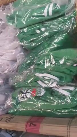 The video shows stacks of green and white t-shirts, most of them individually packaged in clear plastic. The t-shirts have some kind of logo on them, and the green ones appear to have white stripes on the sleeves. The t-shirts are on a wooden pallet. There are also some black t-shirts visible at the bottom of the frame. The scene appears to be a warehouse or storage area. The lighting is somewhat dim.