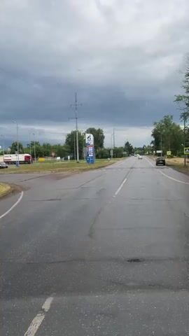 The video shows a road in Zaraysk, Russia, on a cloudy day. The road is wet, indicating that it has recently rained or is still raining. There are power lines overhead and trees lining the side of the road. In the distance, there are some buildings and a large sign for a gas station. The gas station sign displays prices for different types of fuel, including "G-DRIVE" and "GAZPROMNEFT." The prices are displayed in Russian currency, with numbers like "95.09" and "69.51." There are also logos for various payment methods, such as Visa and Mastercard. The overall scene is one of a somewhat overcast and possibly humid day, with the road leading towards a commercial area. There are no people clearly visible in the video, and the focus is on the road and the gas station signage. The time of day appears to be daytime, given the ambient light, but the cloudy sky creates a darker, more subdued atmosphere.