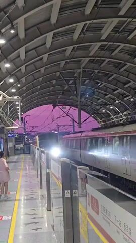 This video captures a metro train arriving at a station in Linping District, China. The setting is a covered metro platform with a distinctive arched, silver-grey roof supported by a network of metal beams. The platform itself is clean and modern, with a yellow safety line demarcated along the edge. A series of sleek, silver metro cars with red and white accents are visible, the leading car of which has its headlights illuminated. The train is in motion, approaching the platform.

In the foreground, a young girl wearing a light pink dress stands near the platform edge, looking towards the approaching train. Another person, also appearing to be a woman, is partially visible further down the platform.

The sky visible through the open sides of the station is a striking, deep pink, suggesting either a very early morning sunrise or a late evening sunset. The lighting within the station is a mix of artificial lights from the ceiling and the train's headlights, casting a bright glow on the platform.

Visible text on signage includes Chinese characters and English translations. One sign displays "下一站 South China," indicating the next stop. Another sign has a warning symbol and Chinese text, likely related to safety precautions. The train itself has the number "019" prominently displayed. The overall atmosphere appears to be one of transit and the routine of urban travel, set against a dramatic and colorful sky.