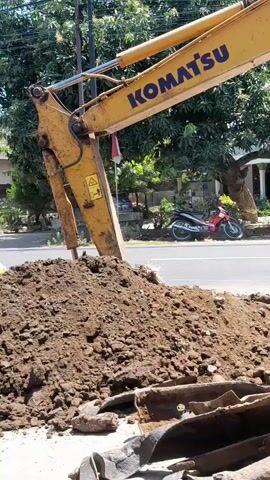 The video shows a Komatsu excavator digging up dirt on the side of a road. The excavator's arm and bucket are visible, as is a pile of excavated dirt. In the background, there are trees, houses, and a street with traffic. A red motorcycle is parked on the side of the road, and a person on a motorcycle passes by in the middle of the video. Another orange car passes by on the road as well. The weather appears to be sunny.